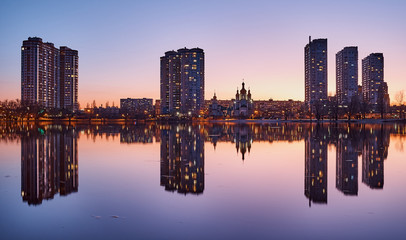 Fototapeta premium Night city on the lake. Reflected buildings. Lilac sunset. Church between the houses. High-rise buildings. Night lights.