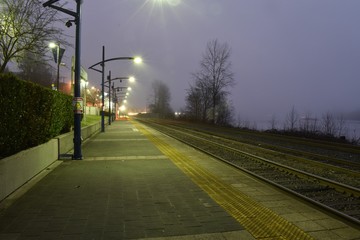 Evening view of deserted outdoor train station. Foggy, overcast.  Tracks beside river. 