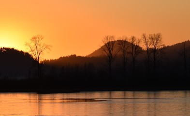 Orange and purple misty sunset, silhouette of trees in front of hills, with calm lake in foreground.  