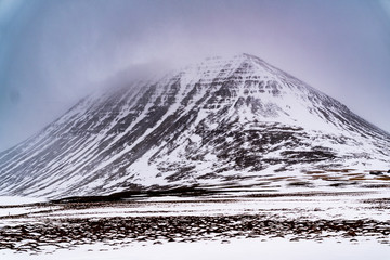 A large pointy mountain raises over the snowy landscape with the cloud covered sun creating an aura of light.