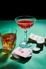 glasses of cognac and cocktail near dollar banknotes, casino tokens and playing cards on green surface isolated on black