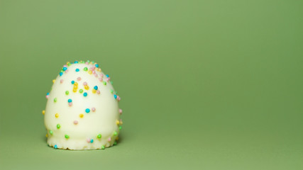 Ein Ei aus weißer Schokolade mti bunten Streuseln aus Zucker isoliert vor grünem Hintergrund, Illustration Ostern, naschen, Süßigkeiten