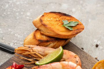 close up view of fried shrimps with grilled toasts and lime on wooden board on grey concrete background