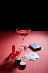 glass of cocktail near playing cards, casino chips and dice on red surface isolated on black