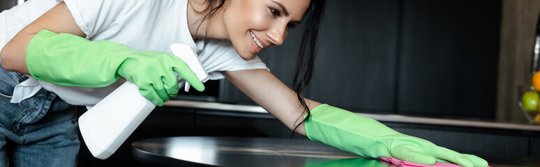 happy girl in latex gloves cleaning table with rag and spray bottle, horizontal crop