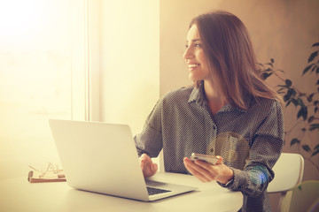 Young woman with smartphone working on laptop at table in cafe