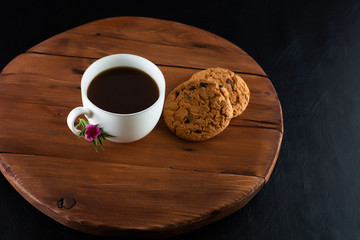 Coffee cup with chocolate cookie on dark wooden background. Cup of coffee with biscuit. Chocolate chip cookies. Black background. Pink flower. Copy space.