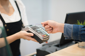 Hand of man customer using wireless or contactless payment of a credit card. Young Asian cashier or seller are smiling to accept payment by nfc technology at retail shop. Contactless payment concept.