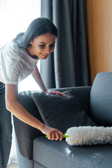 happy beautiful woman cleaning dust from sofa with duster