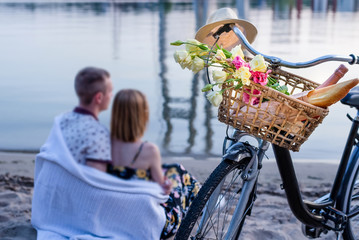 Bicycle basket with flowers,champagne,bread,loaf and hat.Beautiful young couple sitting on the...