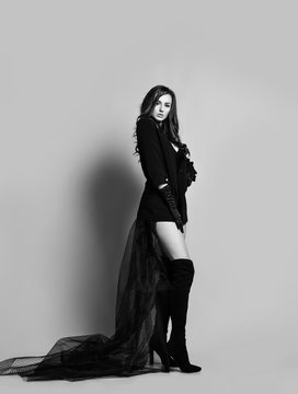 Young beautiful brunette woman in black clothes with long transparent hem and high boots standing and looking at camera