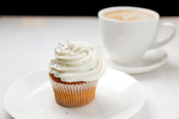 A delicious vanilla cupcake with cream and a cup of cappuccino coffee. Soft focus photo