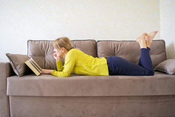 Caucasian woman is reading a book lying at home on the comfortable sofa. Home leisure. It's good to be home.