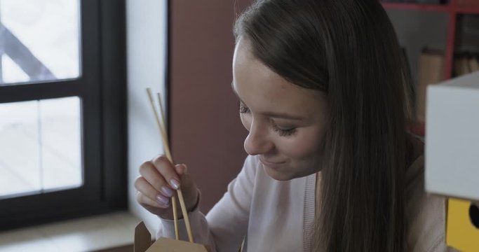Close-up of girl, eat noodles from box with Chinese chopsticks, lunch box, take-away food, food delivery, eats at workplace, smiles, likes noodles, self-isolation mode, take-out food, too lazy to cook