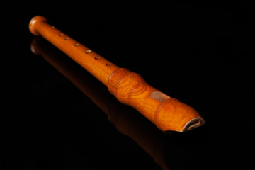 An old wooden flute lies on a black glossy glass, in which its reflection is visible