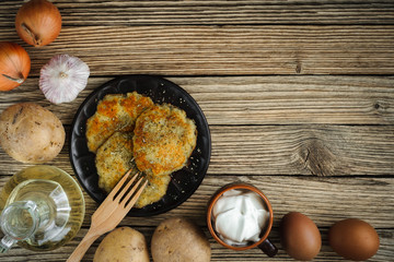 potato pancakes on a clay plate with a wooden fork with raw ingredients on a textured surface from old wooden planks. top view. Artistic rustic mockup for menu, advertisement or blog with copy space
