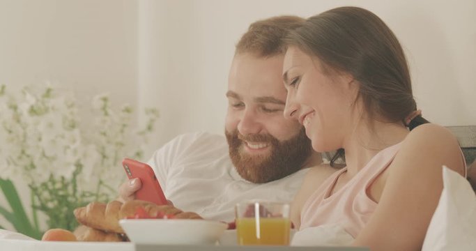 Close up view of couple looking at smartphone screen and smiling while spending time together in bed. Happy girl and man scrolling social media news feed while having breakfast.