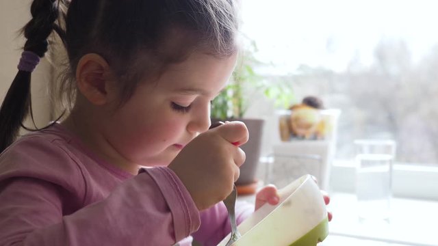 Adorable four years little girl enjoys eating vegetable soup with good appetite, finishing the meal and putting plate on the white table.