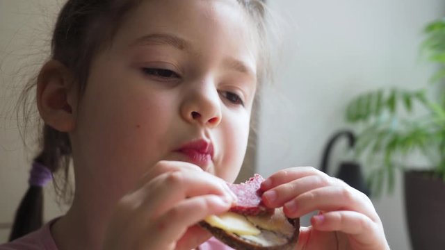 Adorable four years little girl enjoys eating her snack sandwich with good appetite sitting at the white table. Close up