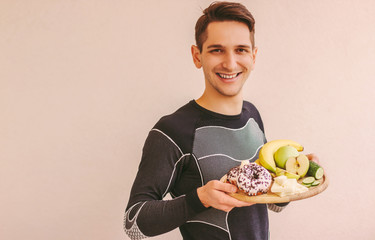 Young sports man deciding what to eat: healthy fruits or sugar junk unhealthy snacks. Happy sports man holding wooden board with fresh apples, bananas, vegetable, donut, chocolate. Dieting concept