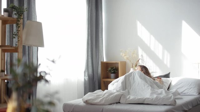 Tracking wide shot of young pretty brunette woman waking up in her bed at home, sitting up and enjoying bright morning sunlight shining through the window