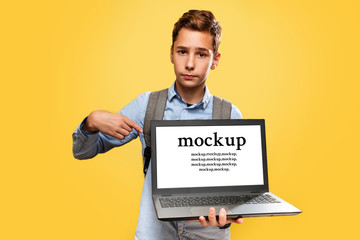 Concept of modern technologies and education. A disgruntled Caucasian teenage boy holds an open laptop and points at it with his finger. Yellow background. Copy space. Mock up