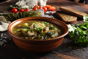 Rustic soup with vegetables, meat and fresh herbs in a clay bowl on an old wooden background. Bread, cumin, coriander, tomatoes, spoons and a bunch of dill. Background image, copy space