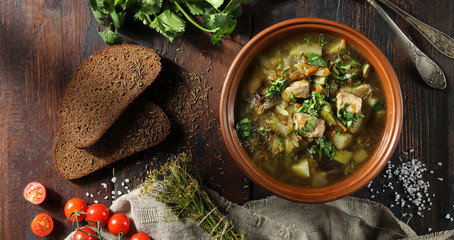 Rustic soup with vegetables, meat and fresh herbs in a clay bowl on an old wooden background. Bread, cumin, coriander, tomatoes, spoons and a bunch of dill. Flatlay. Background image, copy space