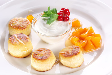 Cheese cakes with sour cream, slices of sweet peach, mint and red currants on a white plate.