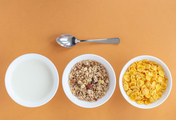 Flat lay with cornflakes, milk and muesli in three white bowls and  metal spoon on beige...
