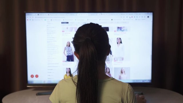 Back view of brunette female, which looks online fashion store on a big display of PC. 
A woman is looking for dresses and other clothes to order online.