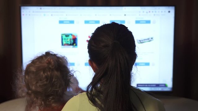 Back view of brunette female and her curly daughter are looking for toys in an online store to order online.