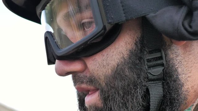 A soldier with black beard in a black helmet and glasses is saying something