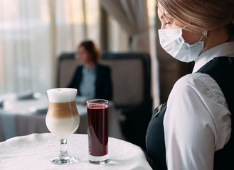 A female Waiter of European appearance in a medical mask serves Latte coffee.