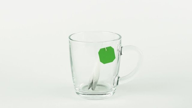 black tea bag with green label is placed in glass transparent mug and filled with boiling water isolated on white background slow motion