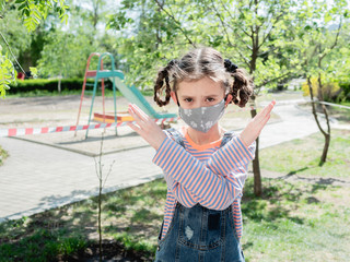 A girl in a protective medical mask on the background of a children's Playground shows a forbidden gesture with her hands. The concept of stay at home.