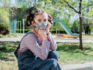 A sad little girl in a protective medical mask sits against the background of a closed Playground during the coronavirus pandemic.