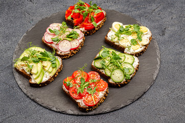 Variety of mini sandwiches with cream cheese and vegetables on plates on a black board, dark background.