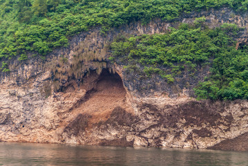 Wuchan, China - May 7, 2010: Dragon Gate Gorge on Daning River. Closeup of cave at water edge in brown rocky cliff with green vegetation above the entrance.