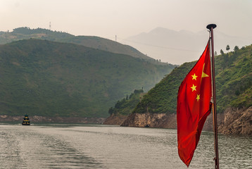 Wuchan, China - May 7, 2010: Dragon Gate Gorge on Daning River. Traditional Chinese sampan boat on green water in green covered mountain canyon with red flag under misty sky.