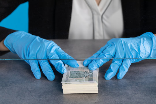 A Young Cashier In A Bank Behind A Glass Holds A Bundle Of Banknotes On The Counter.