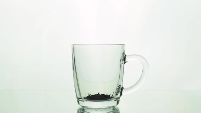 black tea particles pouring into glass transparent mug and adding boiling water to brew tea isolated on white background slow motion