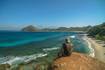 Man with a white cap is sitting at viewpoint camping location next to wild beach and Rinjani Volcano, hills in distance