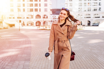 Obraz premium Spring sunny lifestyle fashion portrait of young stylish woman walking on the street, wearing trendy coat drinking coffee