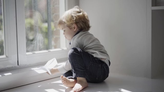 Happy little boy playing with paper plane on white windowsill at home