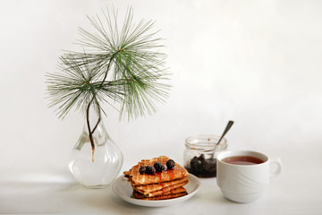 waffles with jam on a white saucer, a Cup of tea on the table and a glass vase with a pine branch