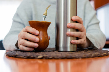 Close-up of middle aged woman's hands holding traditional Argentine yerba mate and thermos of hot water on a wooden table.