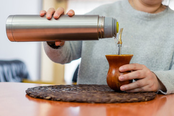 Close-up of middle aged woman's hands serving water from a thermos to traditional Argentine yerba mate in a wooden bowl.