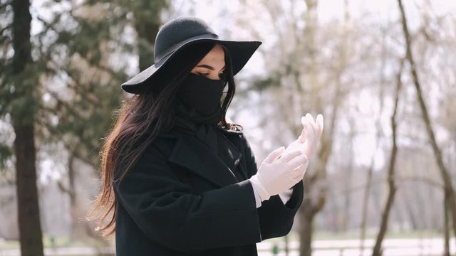 A beautiful young woman in black style is out for a walk. As it is quarantine period so she is wearing a protective mask and gloves in a park.