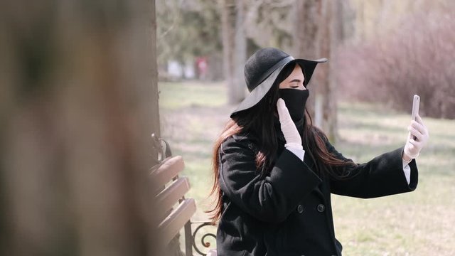 The girl is out for a walk in a park. During quarantine she has to wear disposable protective mask and gloves. She is taking selfies.